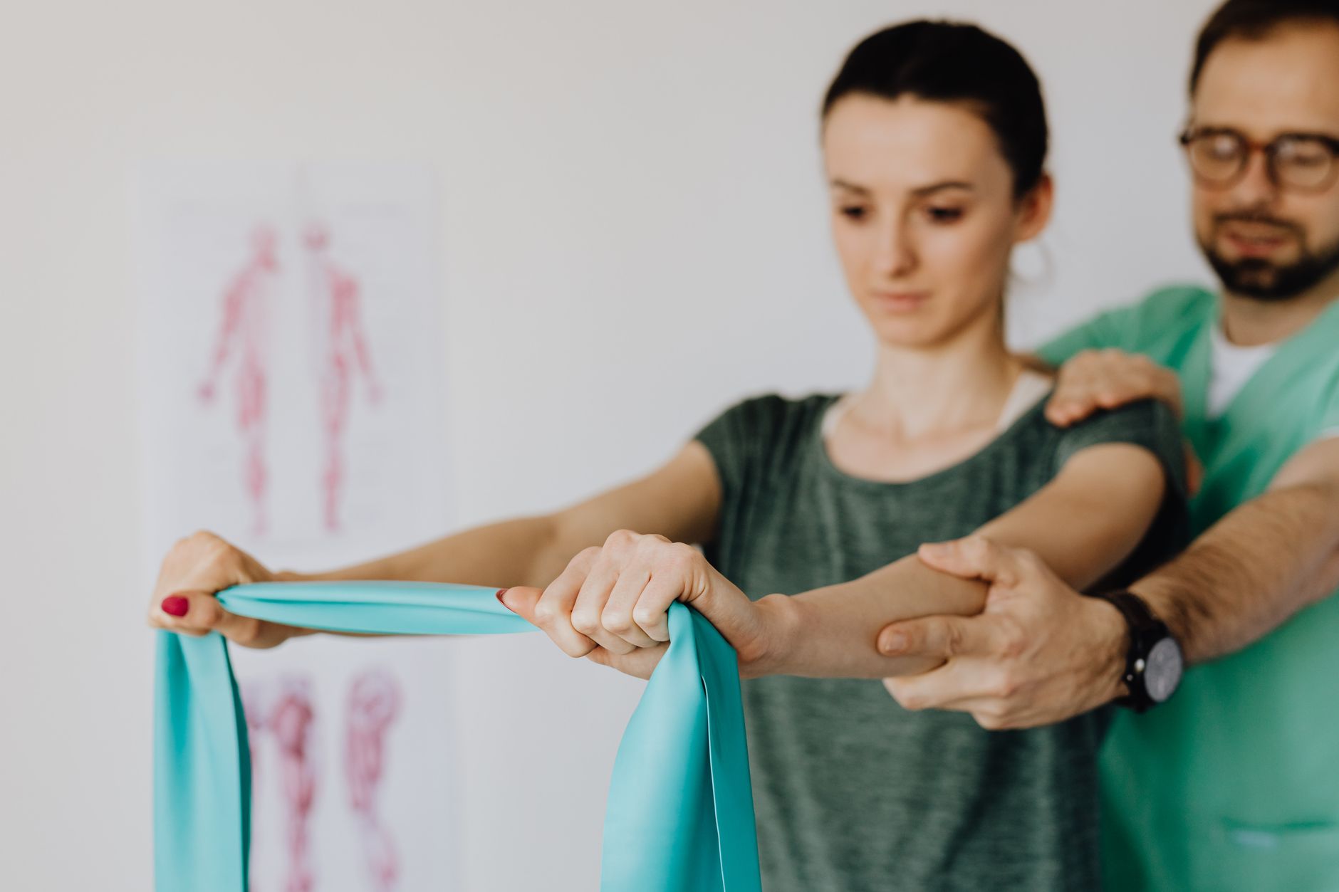 Crop osteopath in uniform and eyeglasses supporting shoulder and reached arm of young woman with elastic tape in doctor office in hospital on blurred background