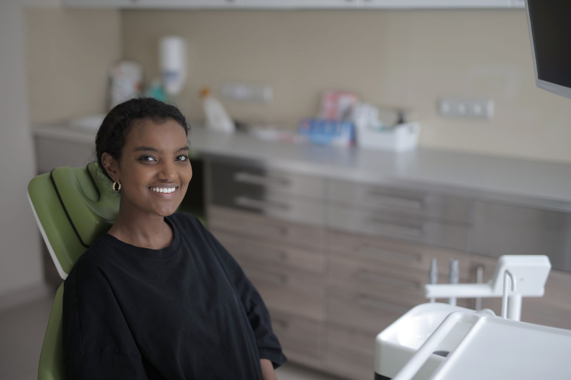 Positive young ethnic female client in black casual clothes smiling while sitting in dental chair and waiting for treatment while looking at camera in modern clinic