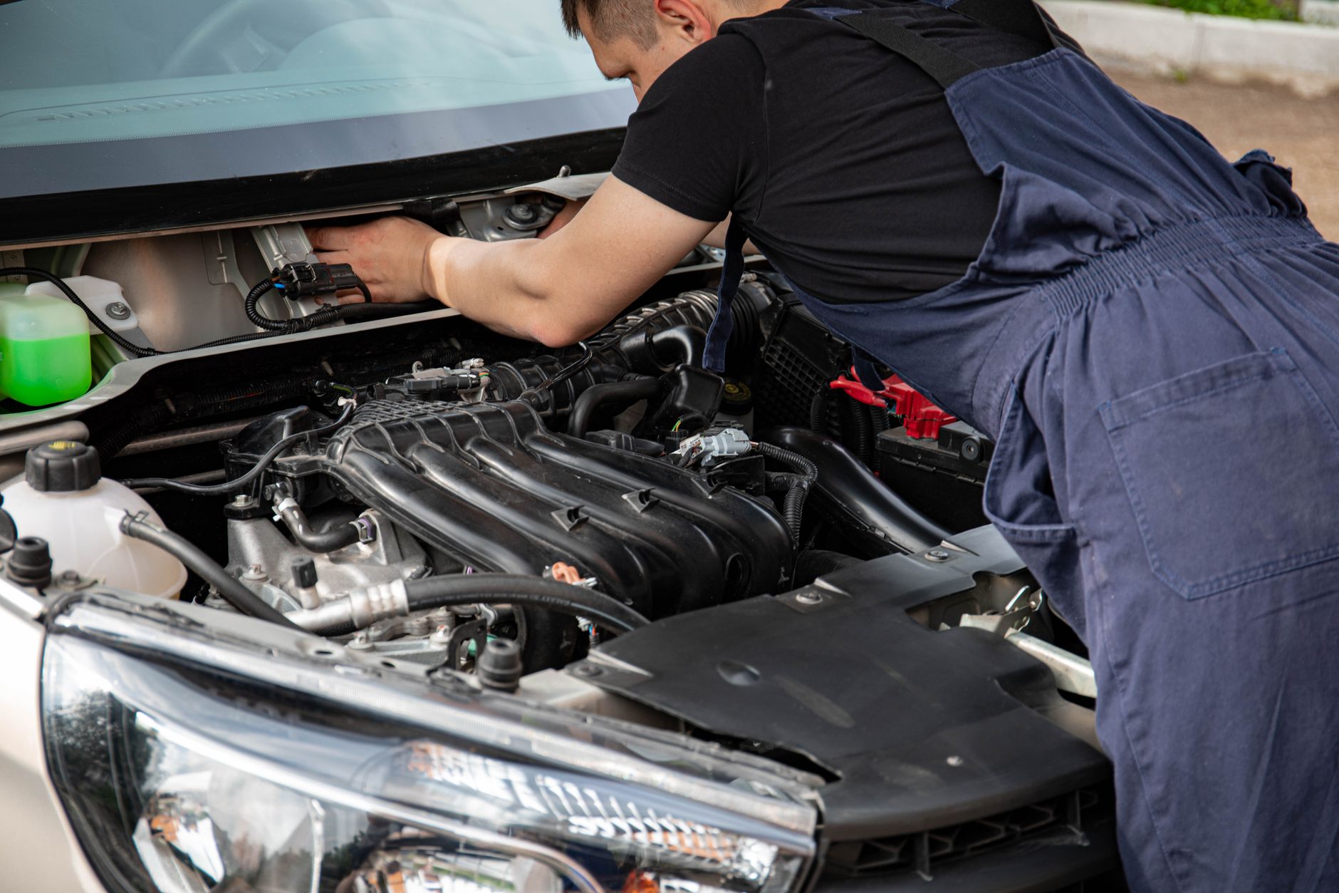 Mechanic in blue overalls repairing a car engine under the hood outdoors.